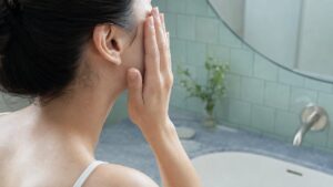 洗面台の鏡の前で、片方の頬に手のひらを添えて化粧水をなじませる女性の後ろ姿（顔は写っていない）