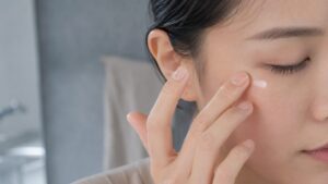 洗面台で薬指の腹を使い、目の下にアイクリームをそっとなじませている女性のクローズアップ