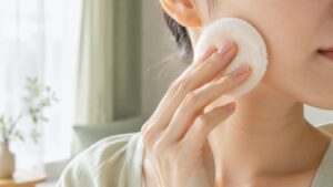 朝の窓辺で、やわらかいコットンパフをそっと頬に置くように触れている女性の手元のクローズアップ